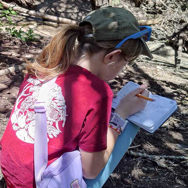 Girl nature explorer outdoors, writing in a Myco Blitz Pocket Field Journal.